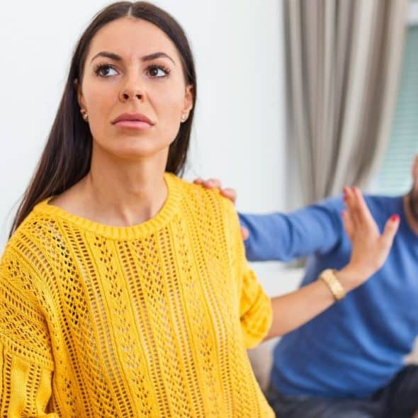 A woman in a yellow sweater looks away while gesturing for a man to stop.