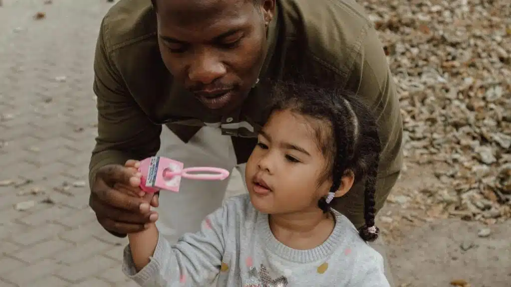 A father gently guiding his child as they play with bubbles outdoors.