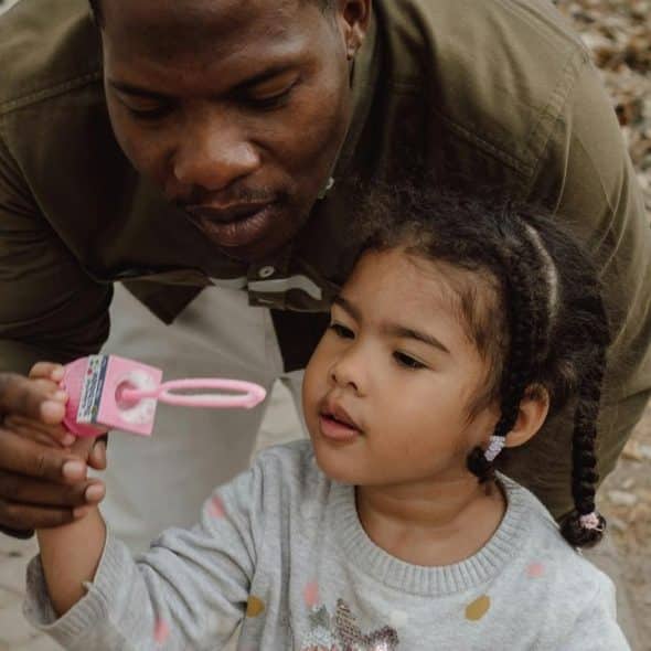 A father gently guiding his child as they play with bubbles outdoors.
