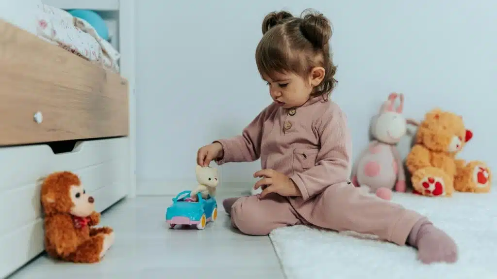 A toddler sitting on the floor playing with a toy car and stuffed animals.