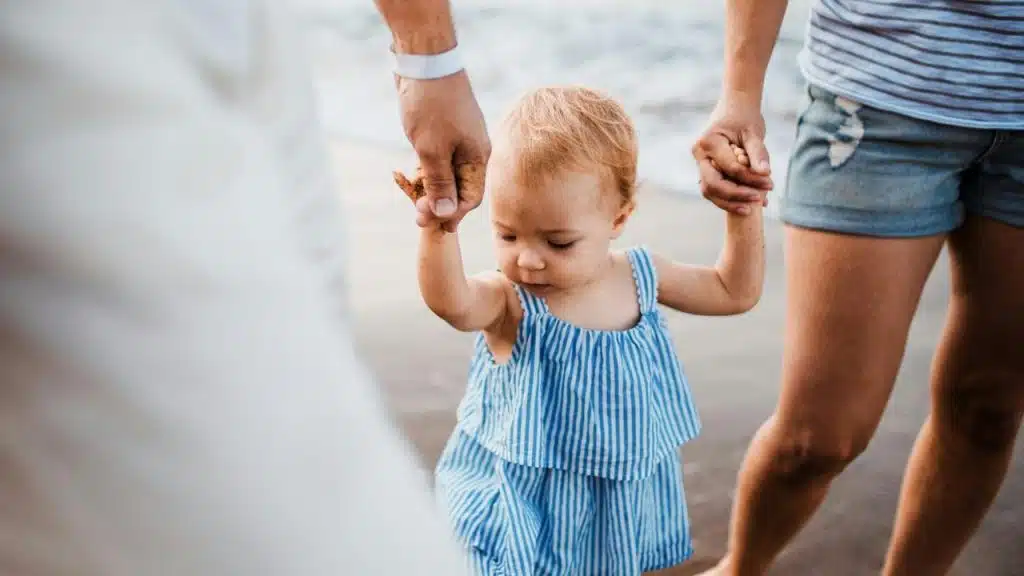 A toddler holding hands with two adults while walking.