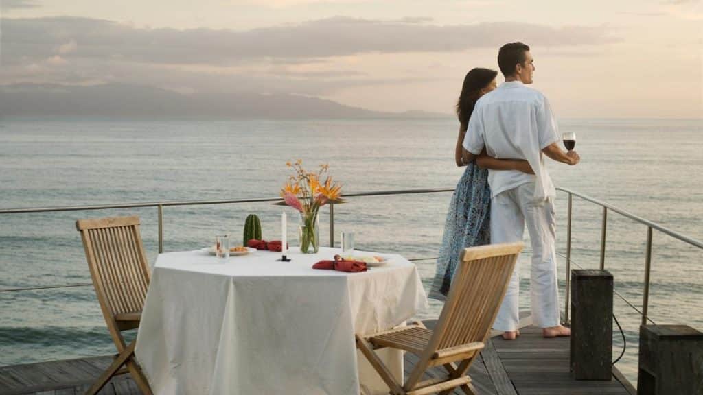 A couple embraces on a seaside deck beside a table set for dinner at sunset.