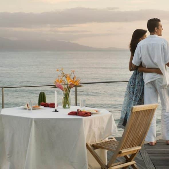 A couple embraces on a seaside deck beside a table set for dinner at sunset.