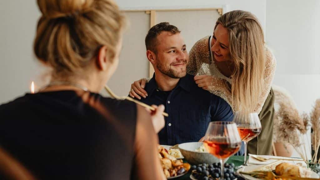 A woman leaning over a man’s shoulder as they smile during a meal with friends.