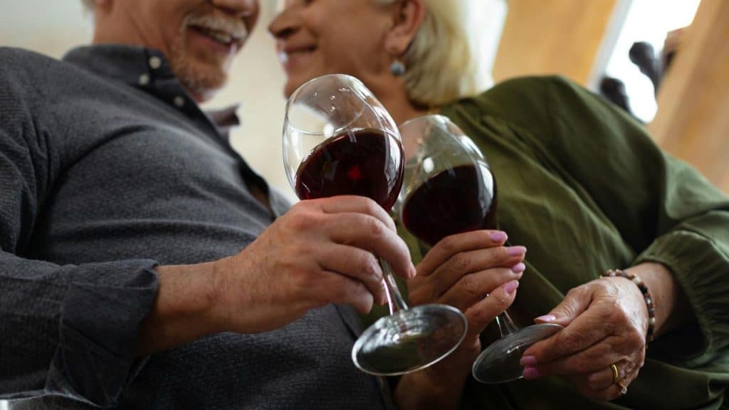 A smiling older couple clinking glasses of red wine indoors.