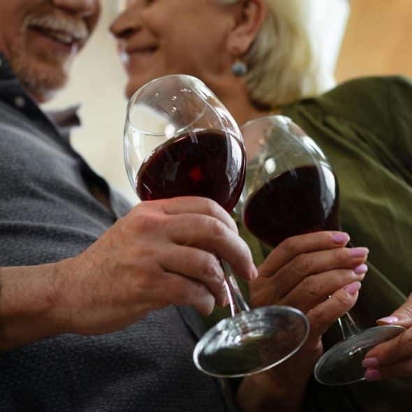 A smiling older couple clinking glasses of red wine indoors.