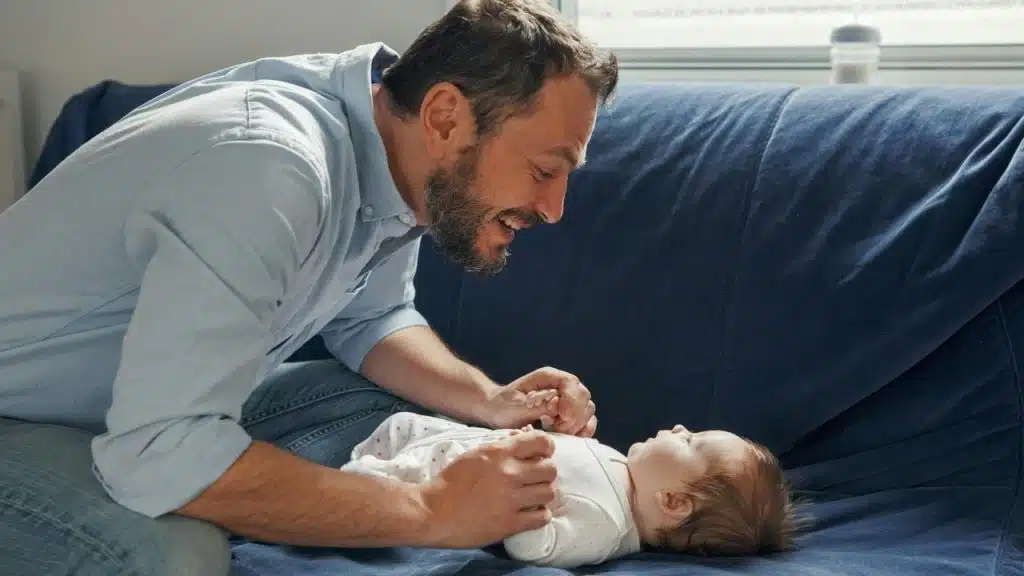 A man smiling and interacting with a baby lying on a couch.