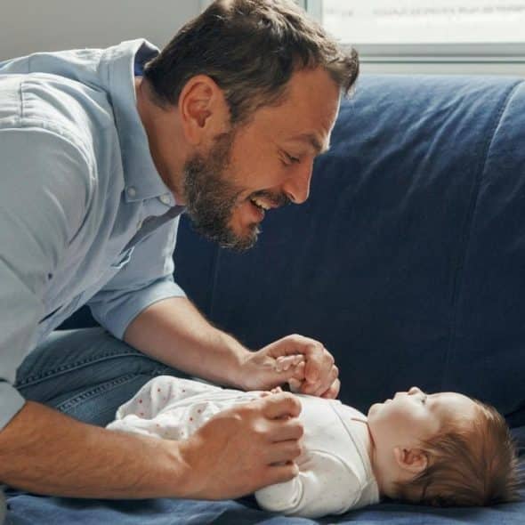 A man smiling and interacting with a baby lying on a couch.
