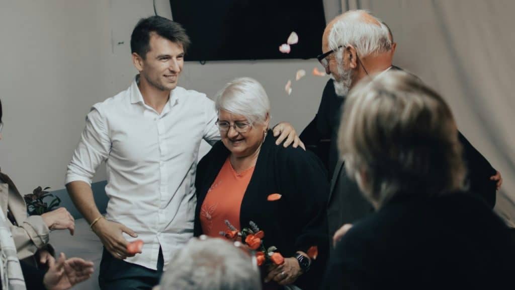 A smiling woman with white hair stands between two men as flower petals fall around them.