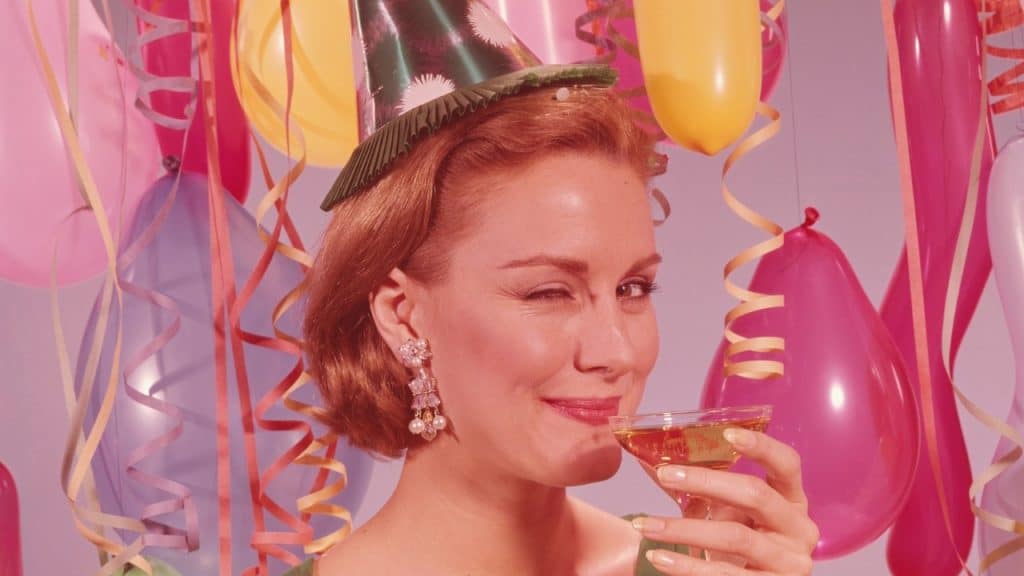 A woman wearing a party hat winks while holding a glass surrounded by balloons and streamers.