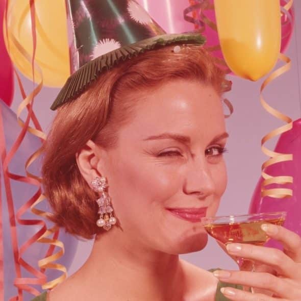 A woman wearing a party hat winks while holding a glass surrounded by balloons and streamers.