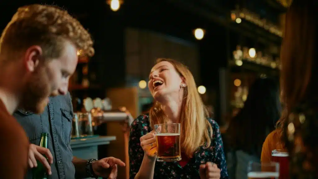 A blonde woman laughs while holding a beer mug in a dimly lit bar setting.