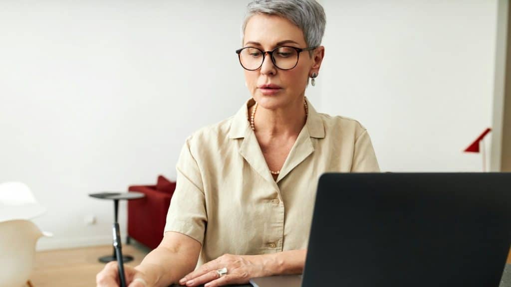 A woman with short grey hair and glasses works on a laptop at a desk.