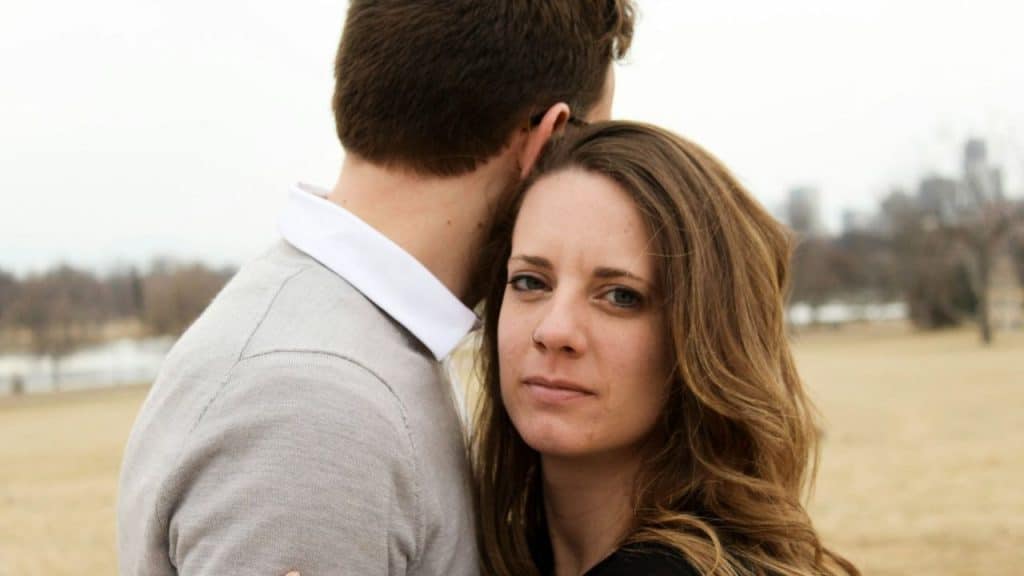 A woman with long brown hair looks forward while standing close behind a man’s shoulder.