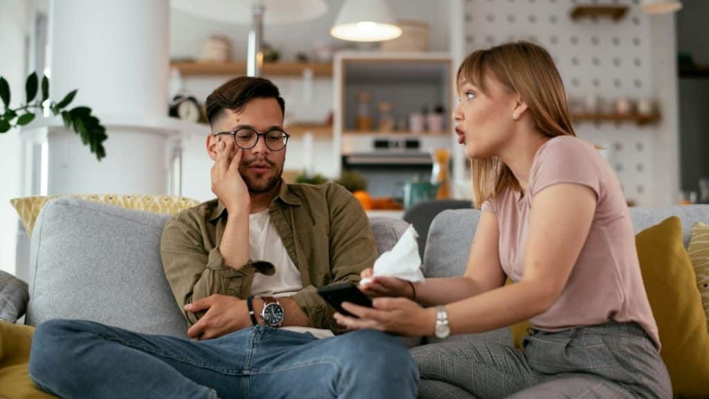 A woman holding a tissue and phone talks to a man sitting on a sofa.