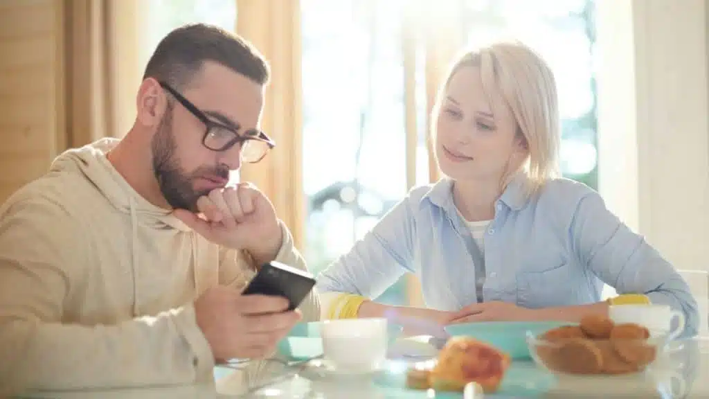 A blonde woman watches a man with glasses and a beard look at his phone.
