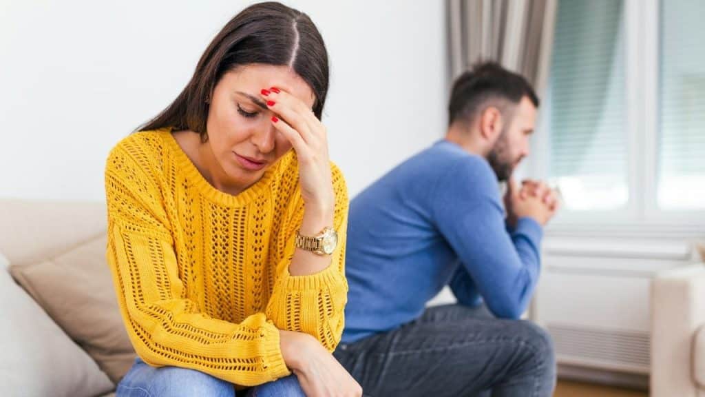 A woman in a yellow sweater holds her head while a man sits behind her.