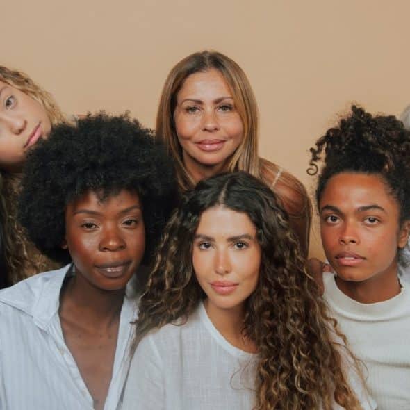 Seven women of various ages and ethnicities pose together against a neutral, tan-colored background.