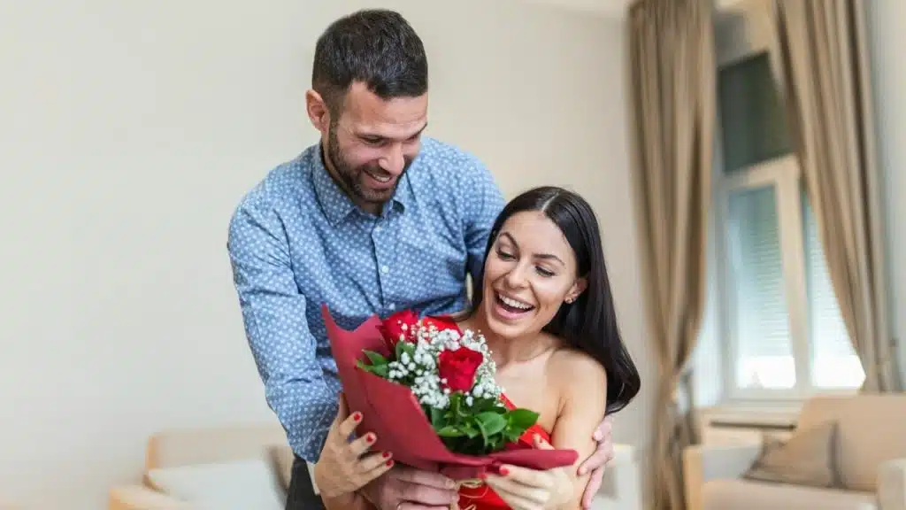A man smilingly presents a bouquet of red roses to a woman who appears surprised and happy.