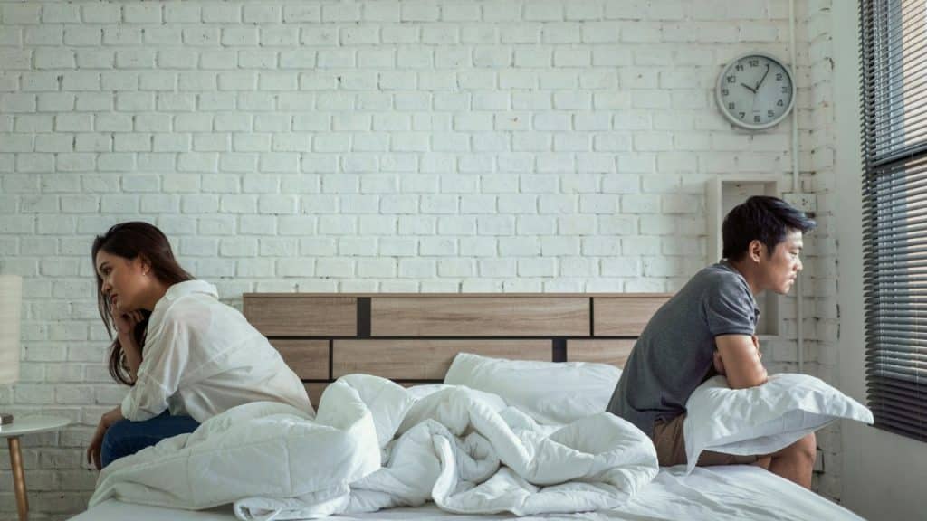 A man and woman sit on opposite sides of a bed, looking away from each other.