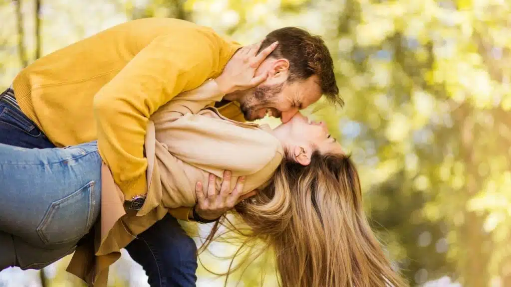 A man in a yellow sweater leans over a woman, holding her as they laugh outdoors.