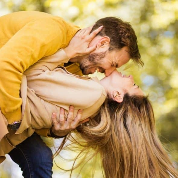 A man in a yellow sweater leans over a woman, holding her as they laugh outdoors.