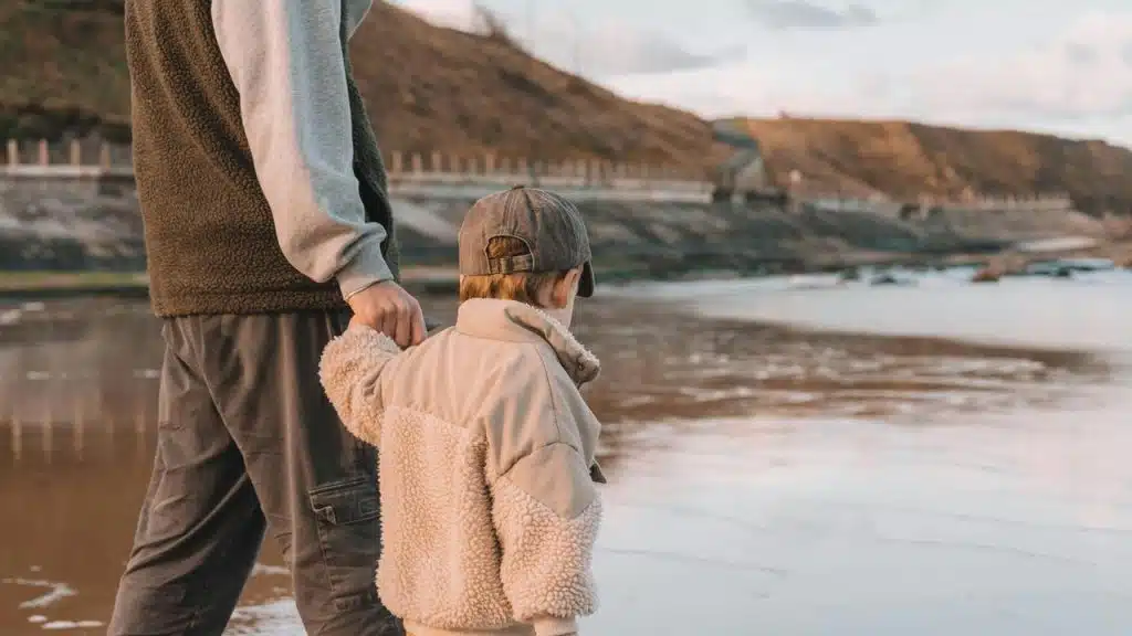 A child holding an adult’s hand while walking along a beach at low tide.