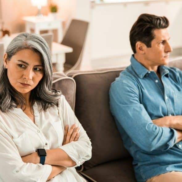 A man and a woman sit on a sofa with arms crossed, looking away from each other.