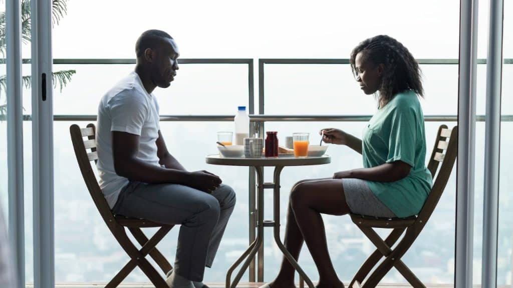 A man and woman sit at a small balcony table eating breakfast in silence.