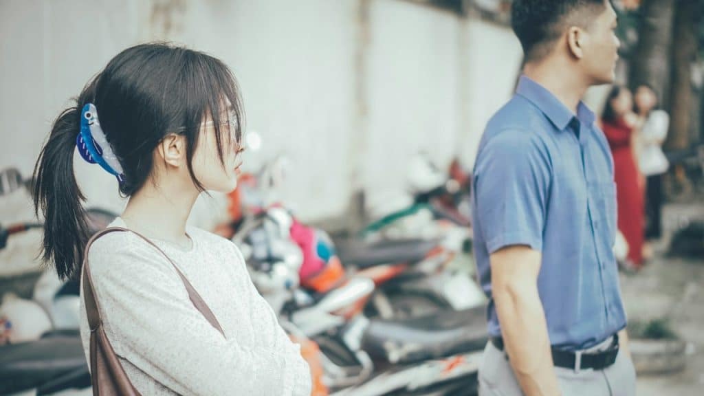 A young woman with arms crossed stands outdoors, looking away from a man.