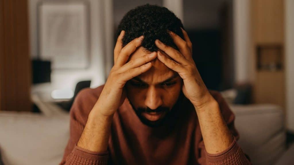A man with dark, curly hair looking down while holding his head with both hands.