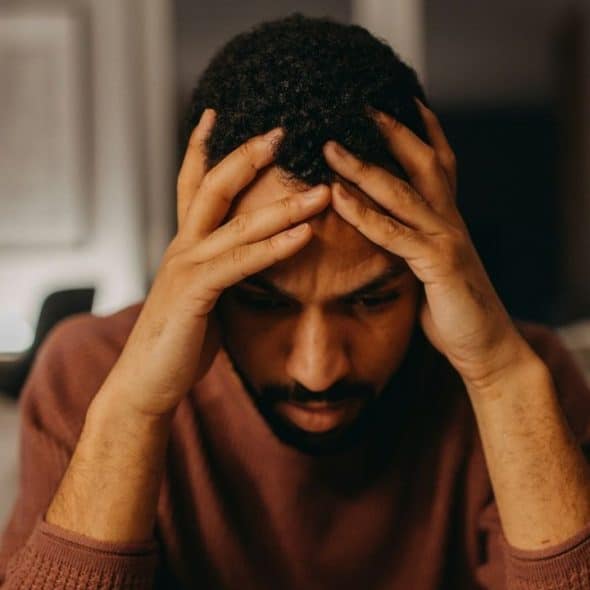 A man with dark, curly hair looking down while holding his head with both hands.