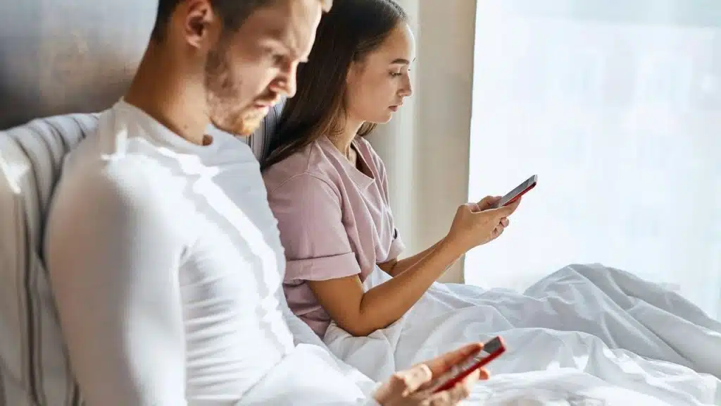 A man and woman sitting upright in a bed, each looking down at their smartphone.