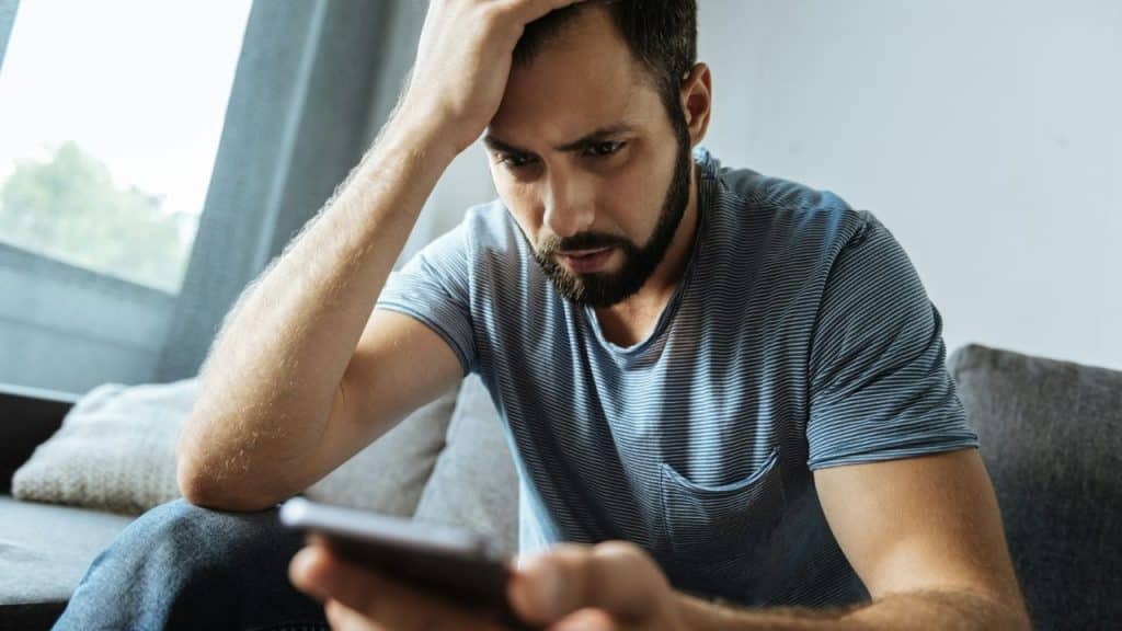 A bearded man sitting on a couch, looking at a smartphone while touching his forehead.
