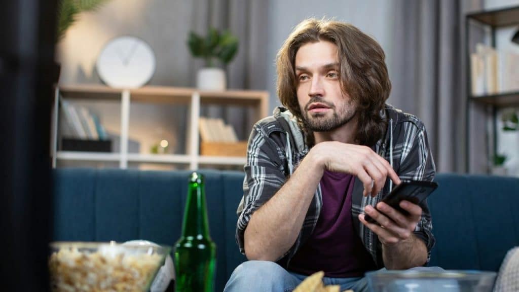 A man with long hair sitting on a couch, holding a phone with snacks nearby.