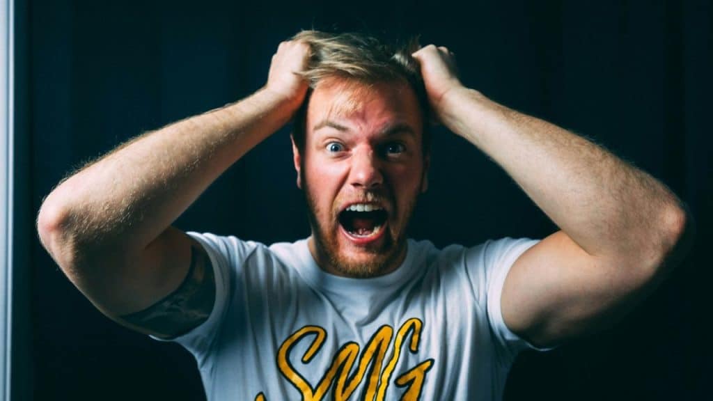 A fair-skinned man with blonde hair shouts with a wide-open mouth while grabbing his hair.