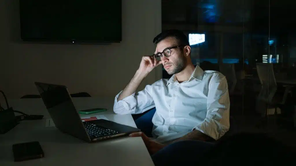 A man wearing glasses sits at a desk in a dark office looking at a laptop.