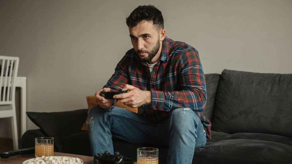A man in a plaid shirt sits on a couch holding a video game controller.