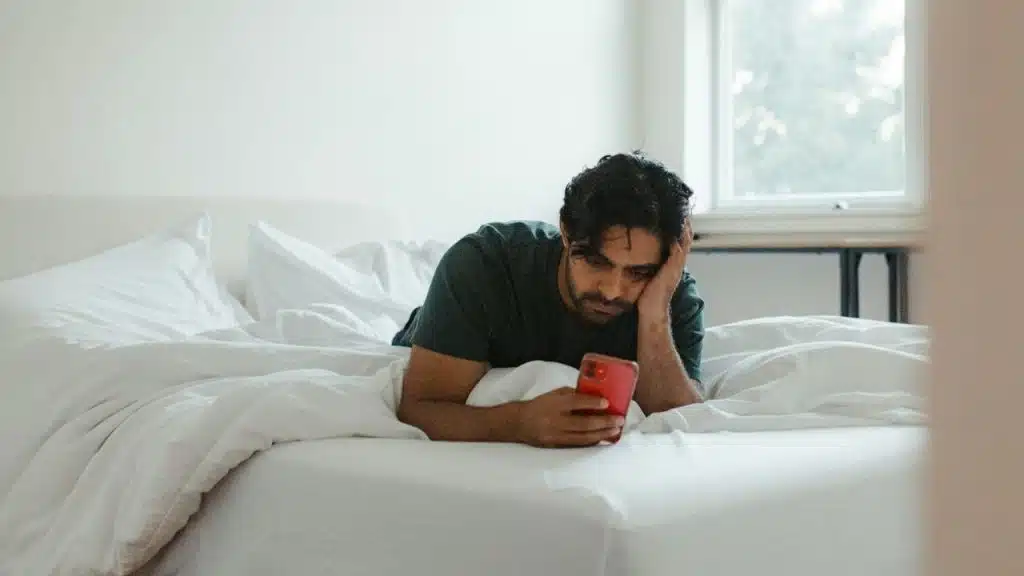 A man lying in a bed looks at a smartphone while propping up his head.