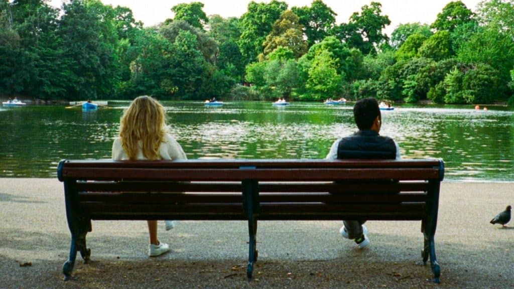 A man and woman sit on opposite ends of a park bench, facing a lake.
