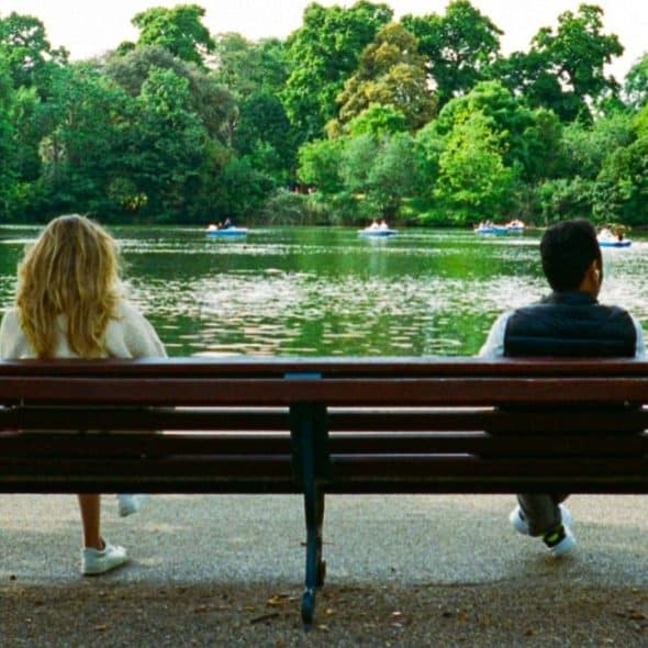 A man and woman sit on opposite ends of a park bench, facing a lake.