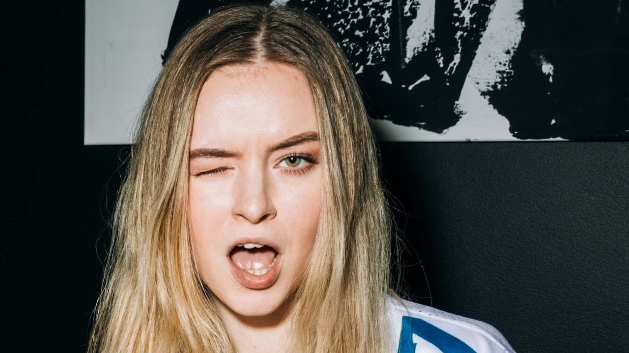A young woman with long blonde hair winks one eye and opens her mouth playfully.
