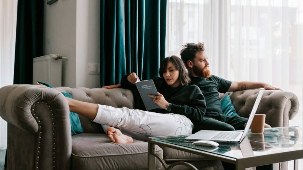 A woman reads a book and a man sits on a sofa near a laptop.