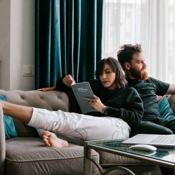 A woman reads a book and a man sits on a sofa near a laptop.