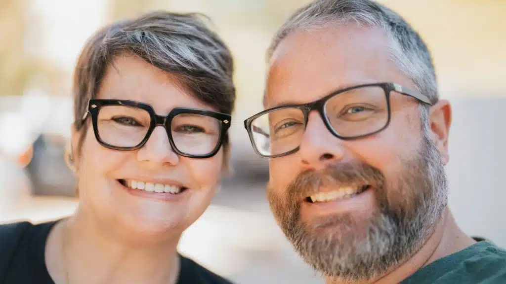 A close-up portrait of a smiling woman and man with glasses outdoors.