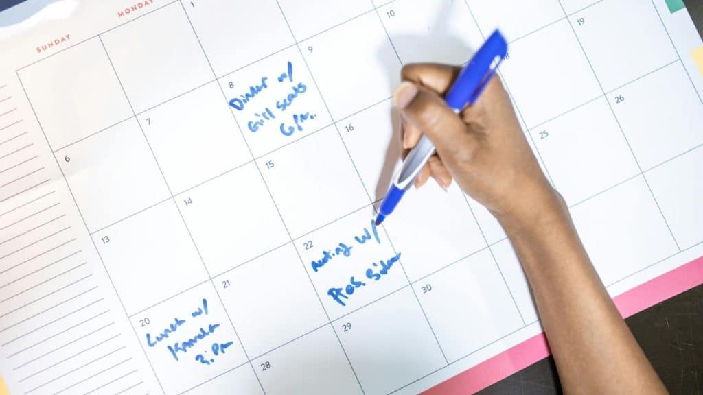 A hand uses a blue pen to write appointments on a large paper wall calendar.
