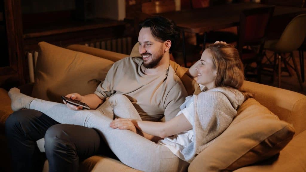 A man and woman sit on a tan couch together, smiling while holding a remote.