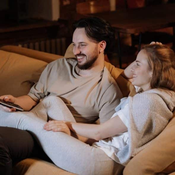 A man and woman sit on a tan couch together, smiling while holding a remote.