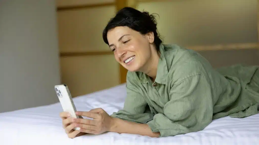 A woman in a green shirt lies on a bed while smiling at her smartphone.