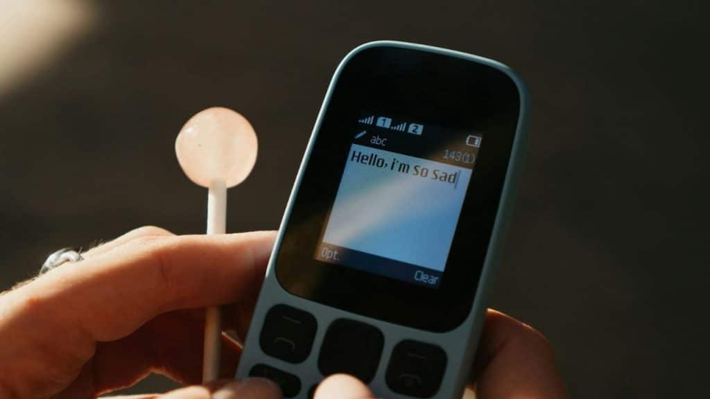 A hand holds a basic mobile phone next to a pink lollipop in bright sunlight.